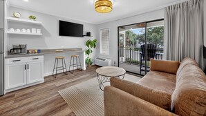 A modern living room with a sofa, a coffee table, and a kitchen area with a TV, shelves, and a plant. - Good Day Sunshine!  Perfect Couples Beach Oasis (Hilton Head Island)