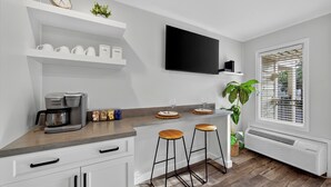 A kitchen with a coffee maker, bar stools, a flat-screen TV, and a window with blinds. - Good Day Sunshine!  Perfect Couples Beach Oasis (Hilton Head Island)