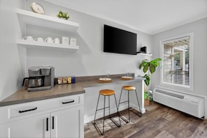 Una cocina con cafetera, taburetes de bar, televisor de pantalla plana y un ventanal con persianas.