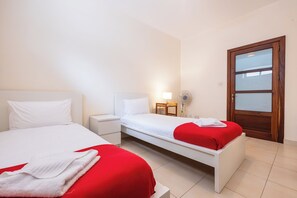 A hotel room with two beds, a bedside table, a lamp, and a door with a frosted glass panel. - budget apartment, affordable stay, cozy apartment, family friendly, group accommodation, patio apartment, outdoor space, free wifi, smart TV, kitchen, central location, value stay, comfortable stay (Il-Mellieħa)