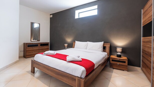 A modern bedroom with a wooden bed, a nightstand, a dresser, and a large window. - budget apartment, affordable stay, cozy apartment, family friendly, group accommodation, patio apartment, outdoor space, free wifi, smart TV, kitchen, central location, value stay, comfortable stay (Il-Mellieħa)