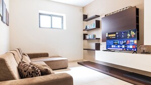 A modern living room with a sofa, a flat-screen TV mounted on the wall, and built-in shelves. - budget apartment, affordable stay, cozy apartment, family friendly, group accommodation, patio apartment, outdoor space, free wifi, smart TV, kitchen, central location, value stay, comfortable stay (Il-Mellieħa)