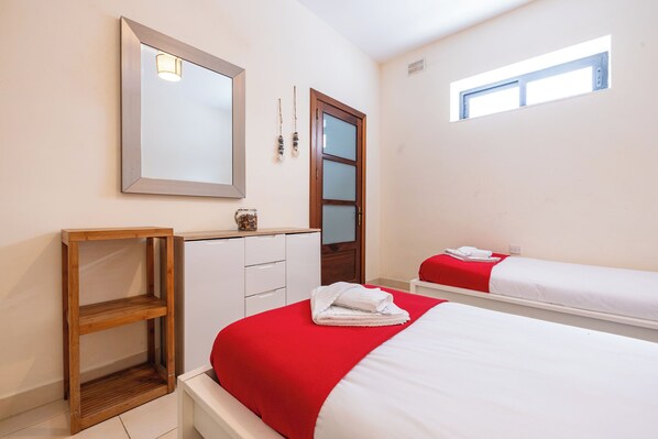 A bedroom with a large mirror, a wooden shelf, a wardrobe, a window, and two beds with red and white bedding. - budget apartment, affordable stay, cozy apartment, family friendly, group accommodation, patio apartment, outdoor space, free wifi, smart TV, kitchen, central location, value stay, comfortable stay (Il-Mellieħa)