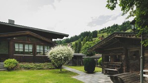 Un bâtiment rustique en bois, doté d’un porche, d’un petit jardin avec un arbre en fleurs et d’un chemin menant à l’entrée.