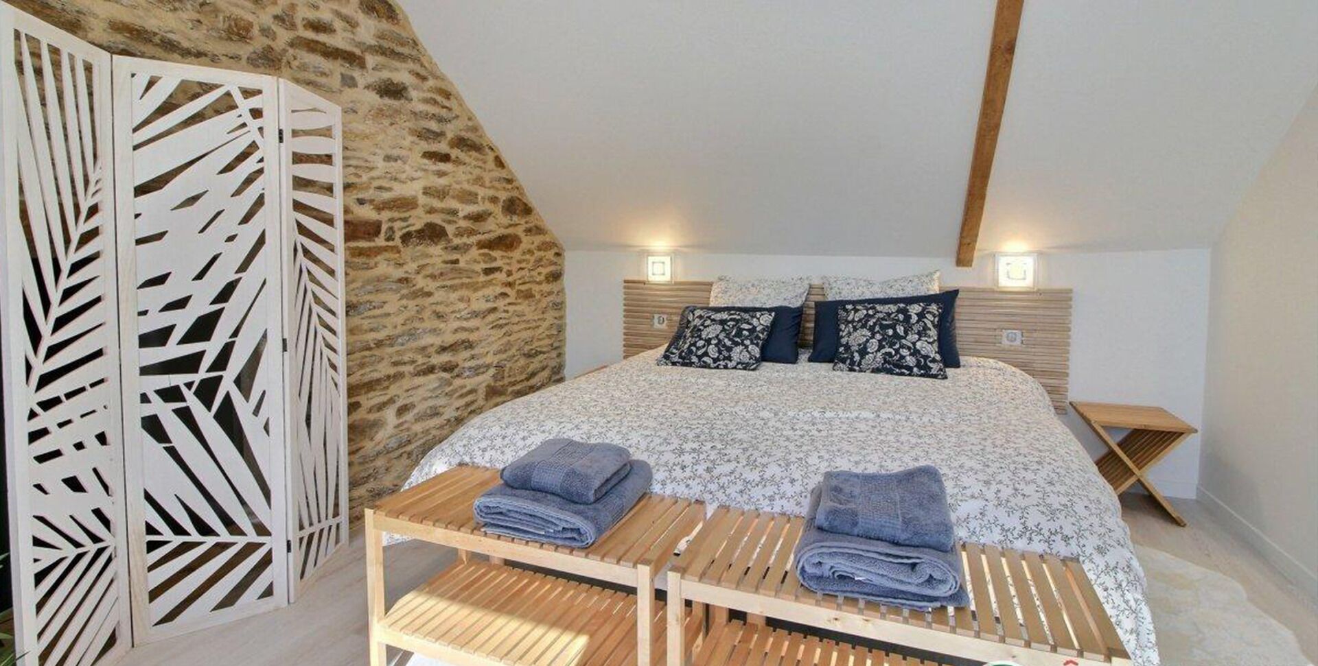 Un dormitorio con techo inclinado, una cama con colcha floral, dos almohadas, un banco de madera con toallas y un separador decorativo blanco.