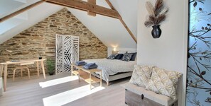 Ein Schlafzimmer mit schrägen Wänden, einer Steinwand, einem Bett mit gemusterten Textilien, einer Couch, einem Schreibtisch und einer Zierpflanze.