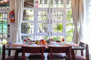 A dining table set with plates, glasses, and fruits, with a fire extinguisher on the wall and a window with curtains. - Alaya Stays Farsauli 1 Pet friendly 3BHK (Nagri Gaon)