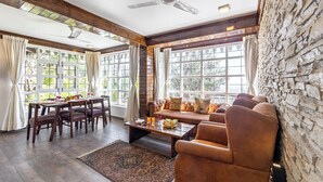 A spacious living area with a dining table, sofa, and chairs, large windows, and a stone wall. - Alaya Stays Farsauli 1 Pet friendly 3BHK (Nagri Gaon)