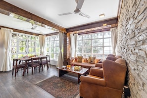 A spacious living area with a dining table, sofa, and chairs, large windows, and a stone wall. - Alaya Stays Farsauli 1 Pet friendly 3BHK (Nagri Gaon)