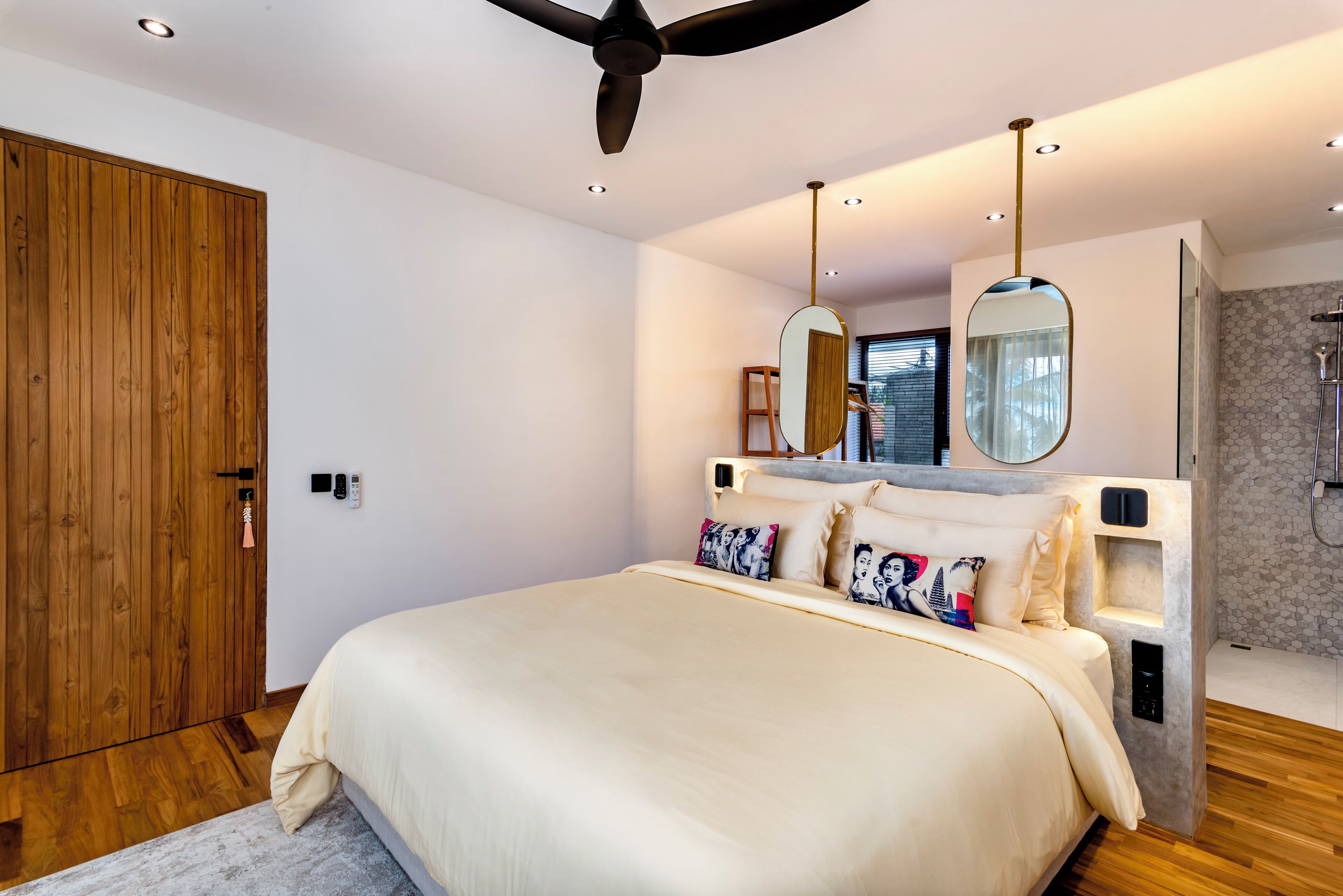 A modern bedroom with a large bed, wooden wardrobe, ceiling fan, and a shower area.