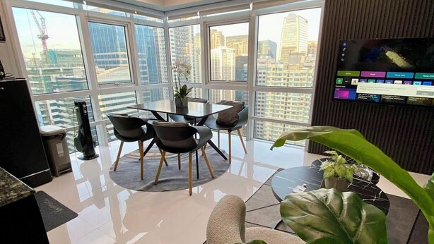 A modern living room with a dining area, large windows, a flat-screen TV, and a large plant. - Hive Loft 2BR BGC w/ Free Parking + Skyline View (Taguig)