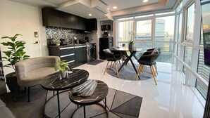 A modern living room with a sofa, coffee table, and chairs, a kitchen area with a tiled backsplash, and large windows offering a city view. - Hive Loft 2BR BGC w/ Free Parking + Skyline View (Taguig)