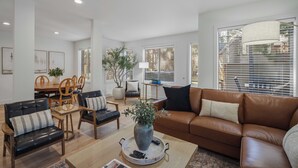 Een ruime woonkamer met een bruine leren bank, zwarte leren fauteuils, een houten eettafel en een potplant.