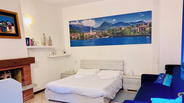 Een slaapkamer met een bed, een nachtkastje, een lamp, een blauwe bank, een open haard en een schilderij van een kustplaatsje.