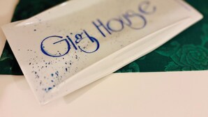 Een wit rechthoekig object met 'Gigi House' in blauw erop, op een groen oppervlak met patroon.