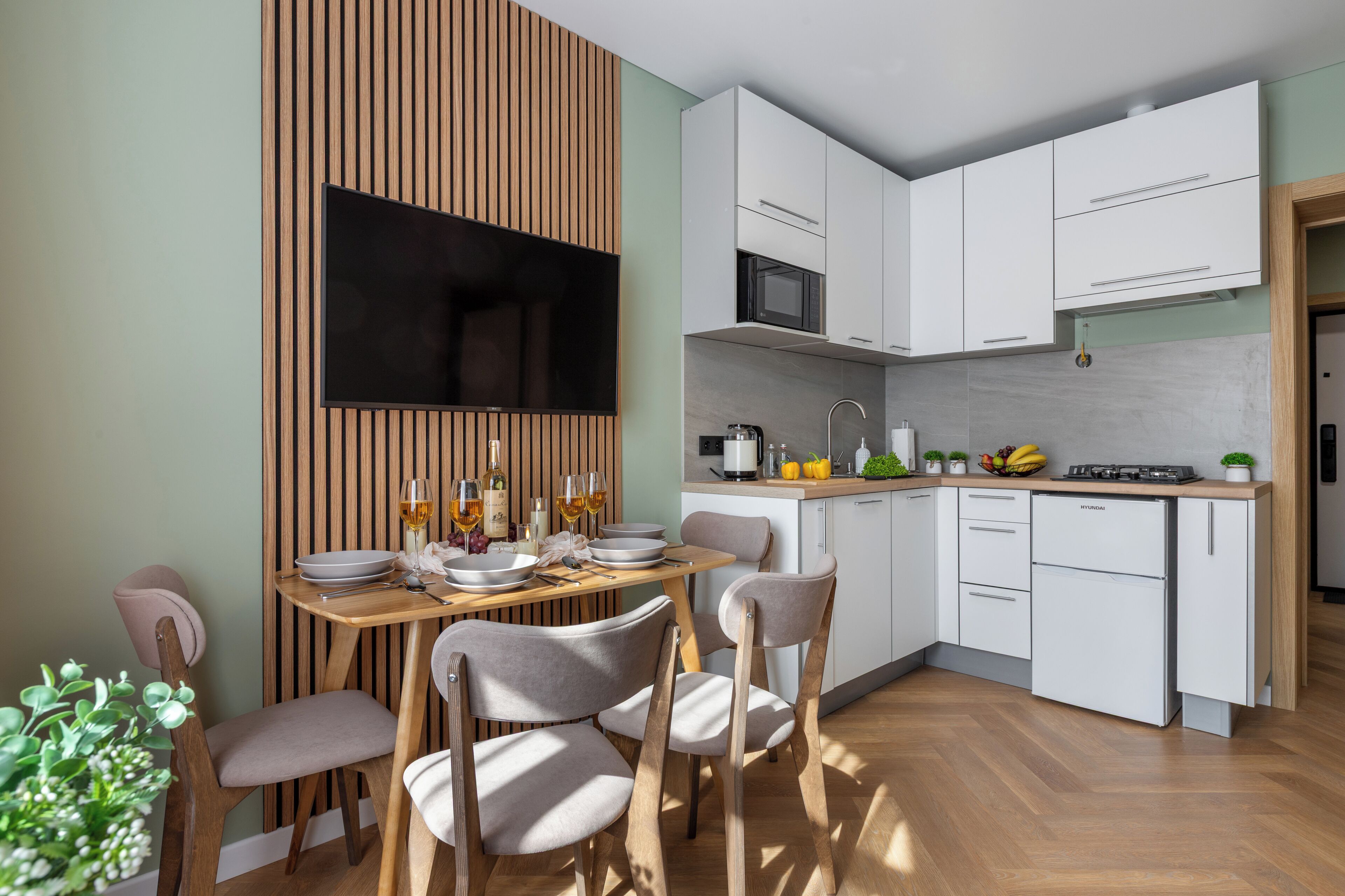 A modern kitchen with white cabinets, a wooden dining table, and a flat-screen TV mounted on a wooden panel.