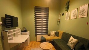 A living room with a TV, a sofa, a coffee table, and a window with blinds. - Roza Mitra Butik Otel (İstanbul)