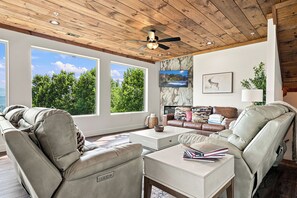 Ein Wohnzimmer mit Deckenventilator, großen Fenstern, einem Kamin und einem an der Wand befestigten Fernseher.