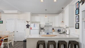 A kitchen with white cabinets, a central island with stools, a microwave, and a refrigerator. - Walk to Beach Arcade Games Family Friendly Oak Island (Oak Island)