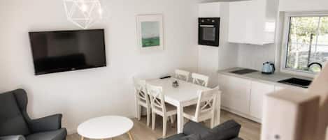 A modern, compact living space with a flat-screen TV, a dining area, a kitchenette, and a sofa.