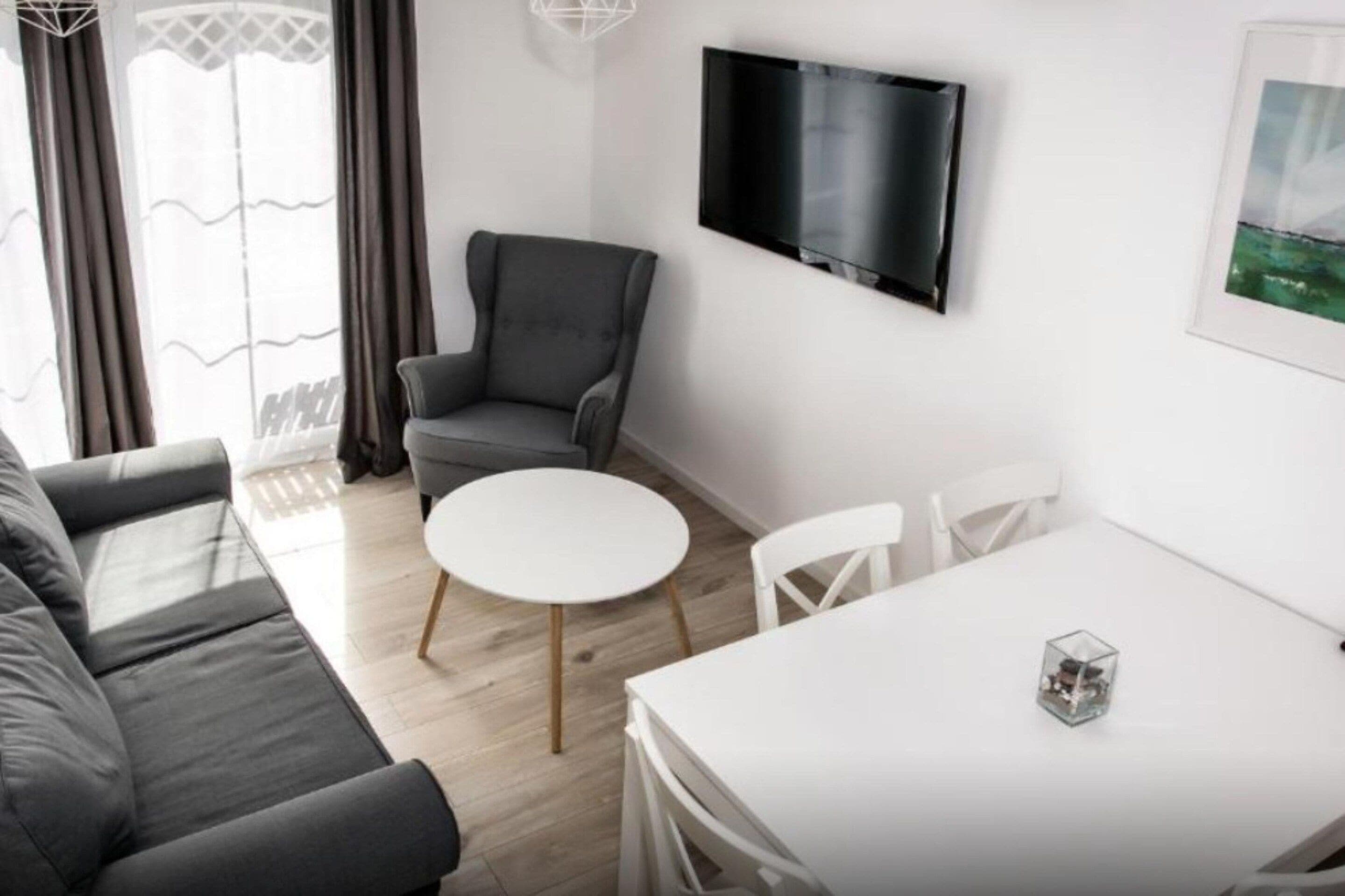 A modern living room with a grey sofa, a round white table, and a grey armchair. A flat-screen TV is mounted on the wall.