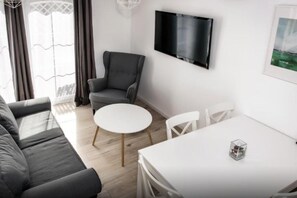 Ein modernes Wohnzimmer mit einem grauen Sofa, einem runden weißen Tisch und einem grauen Sessel. Ein Flachbildfernseher ist an der Wand angebracht.