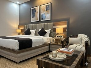 A modern hotel room with a large bed, bedside lamps, a coffee table with a book and candle, and a chair with a blanket. - Lotus Infinity Hotel Haridwar by Akshay (Haridwar)