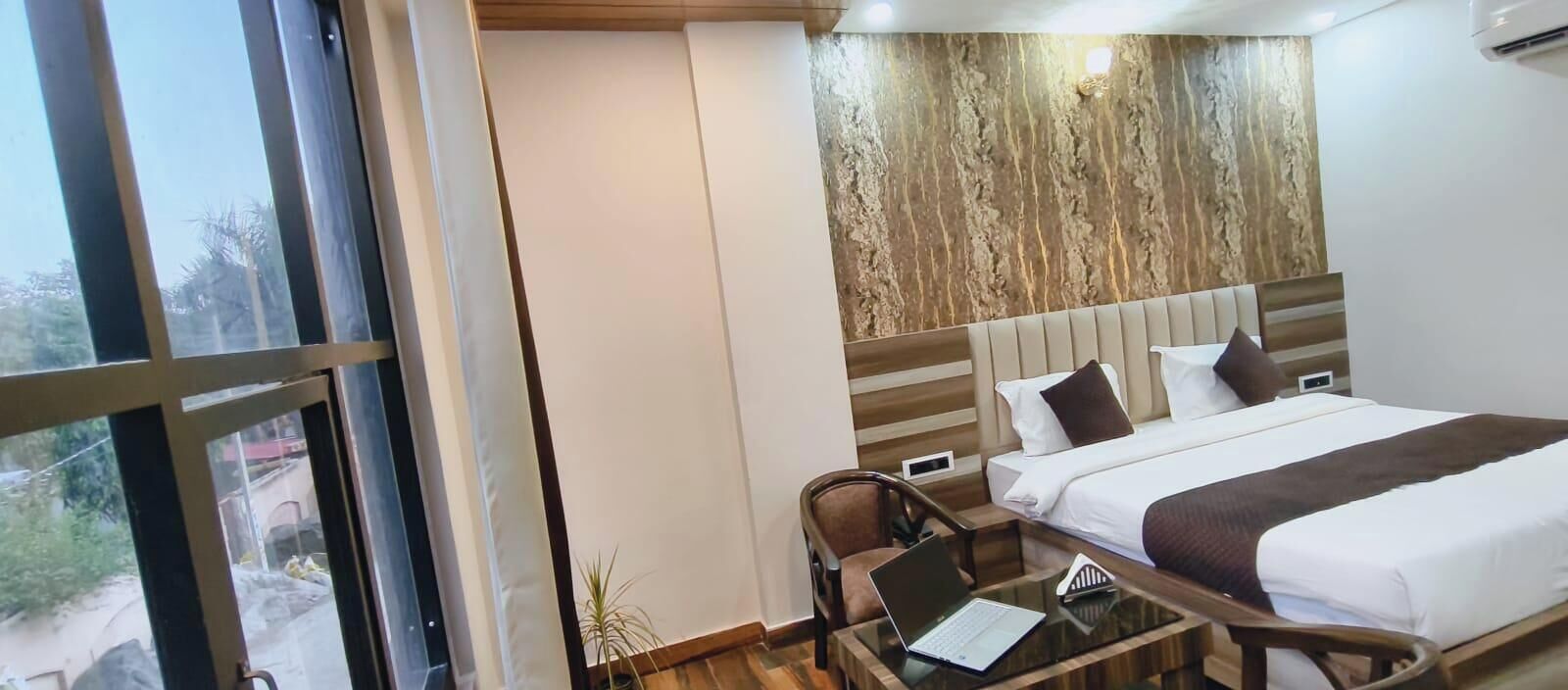 Habitación de hotel con cama, escritorio, computadora portátil, silla y vistas al exterior por la ventana.