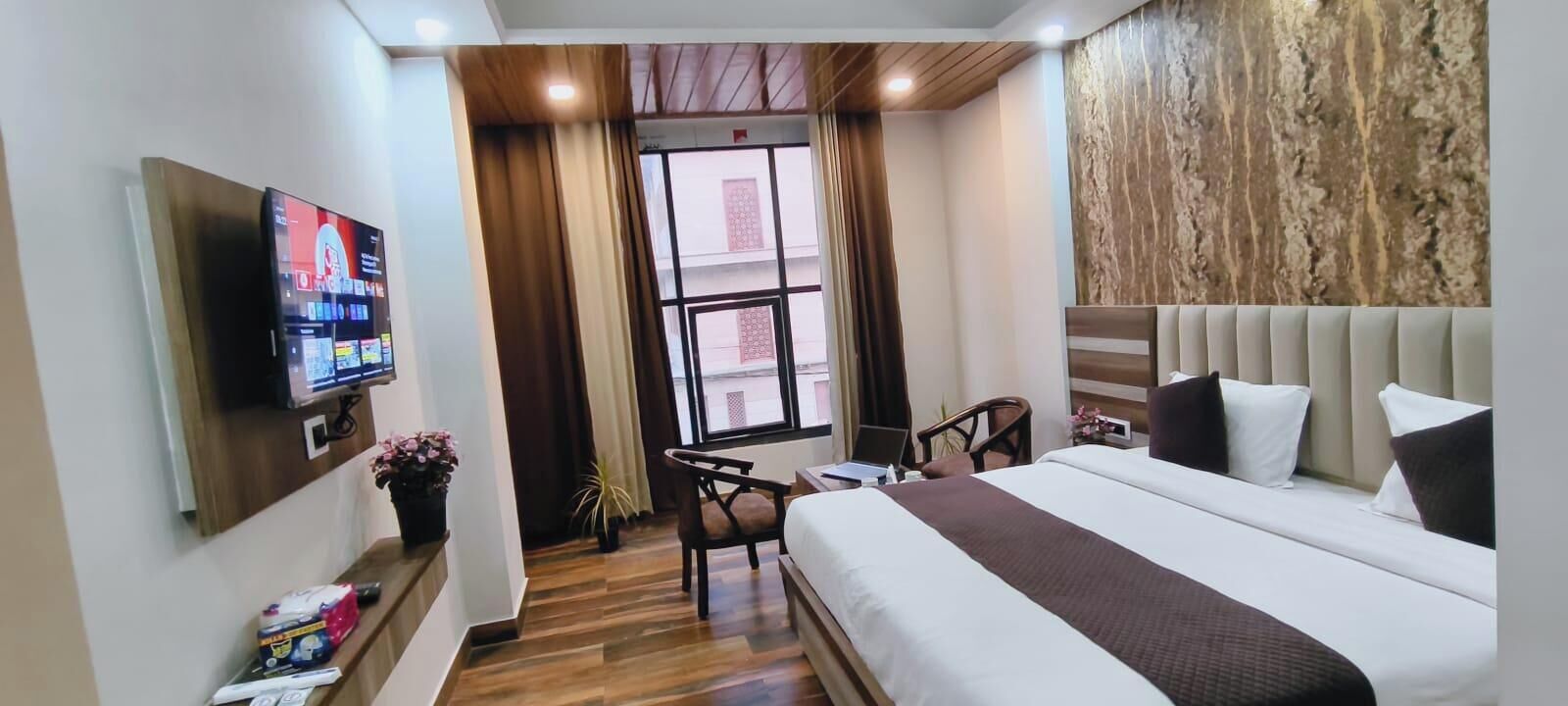 A hotel room with a bed, TV, wooden floor, curtains, and a window.