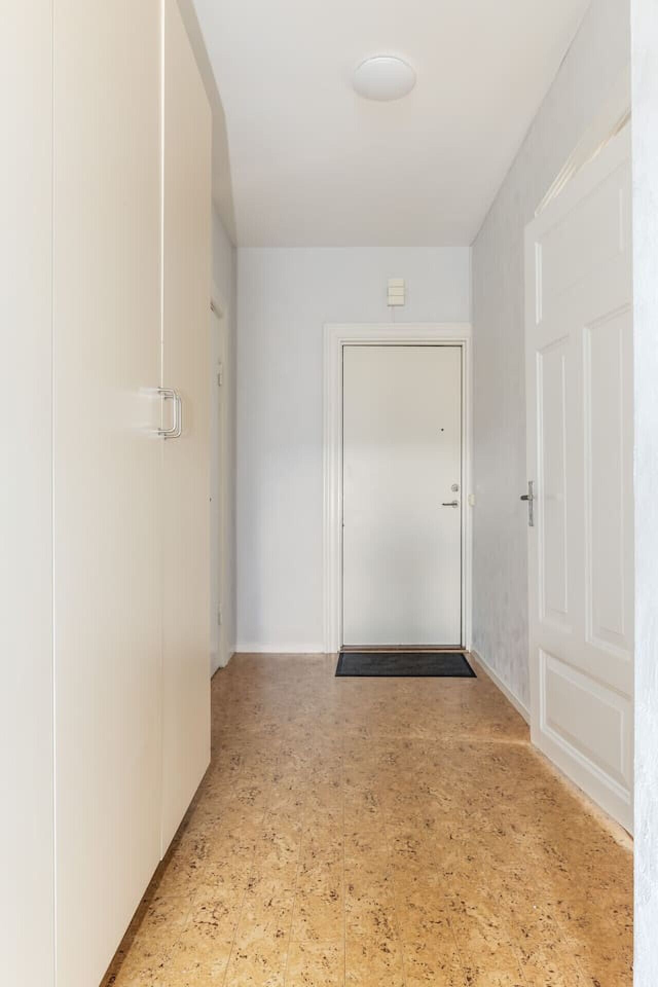Un couloir étroit aux murs blancs et au sol à motifs, qui mène à une porte fermée sur laquelle est posé un paillasson.