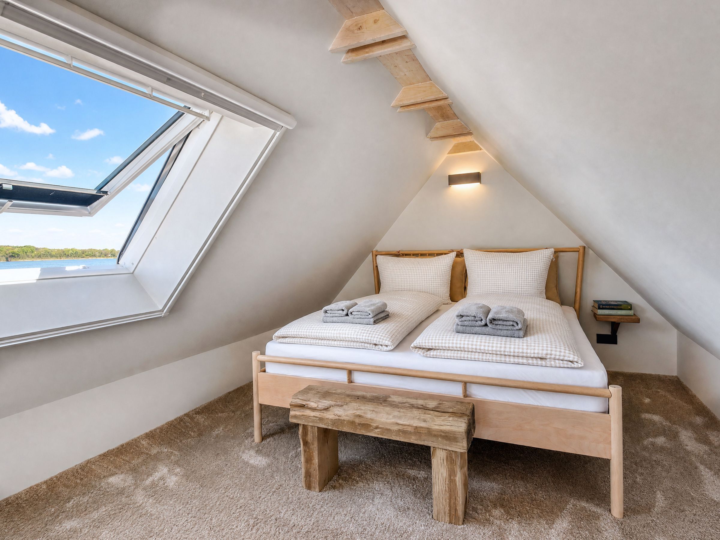 A bedroom with a skylight, a bed with pillows, a bench, and a nightstand with books.