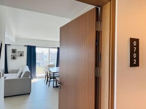Ein modernes Wohnzimmer mit einer Couch, einem Esstisch und Stühlen. Eine Holztür mit Klingelnummer ist sichtbar.