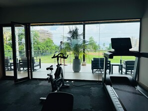 Un gimnasio con equipo de ejercicio, una cinta de correr y un amplio ventanal con vista a un parque y edificios.