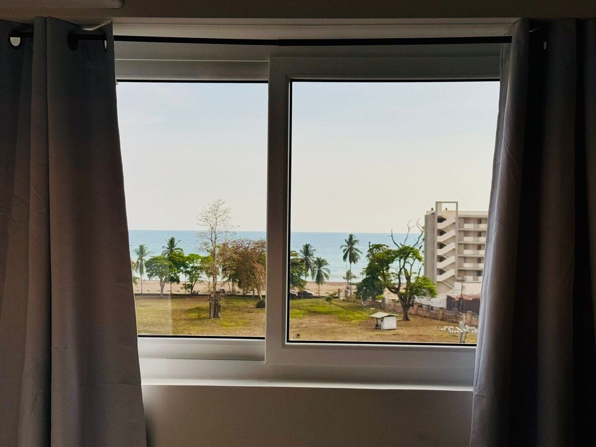 Janela com vista para uma praia, palmeiras e um edifício.