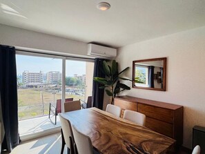 Ein Hotelzimmer mit Esstisch, Stühlen, einem Schrank, einem großen Fenster mit Blick auf Gebäude und einer Klimaanlage.
