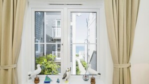 Ein Fenster mit weißen Vorhängen, Blick auf ein Gebäude und zwei weiße Vogel-Figuren auf der Fensterbank.