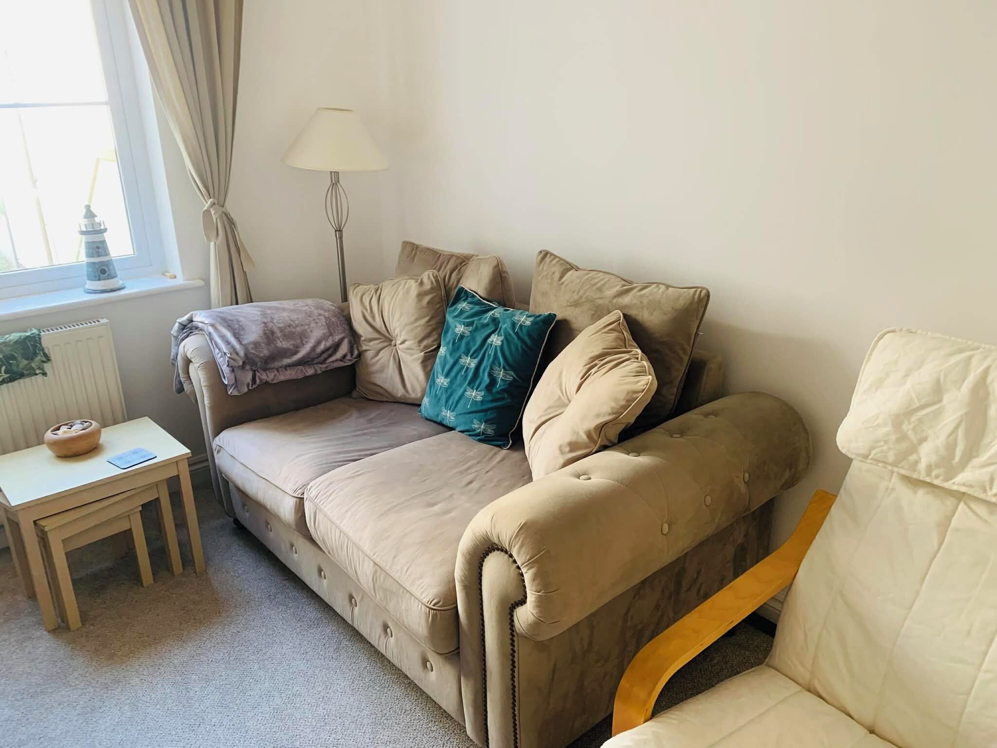 A beige sofa with a cushion, a small wooden table with a bowl, a lamp, and a window with curtains.