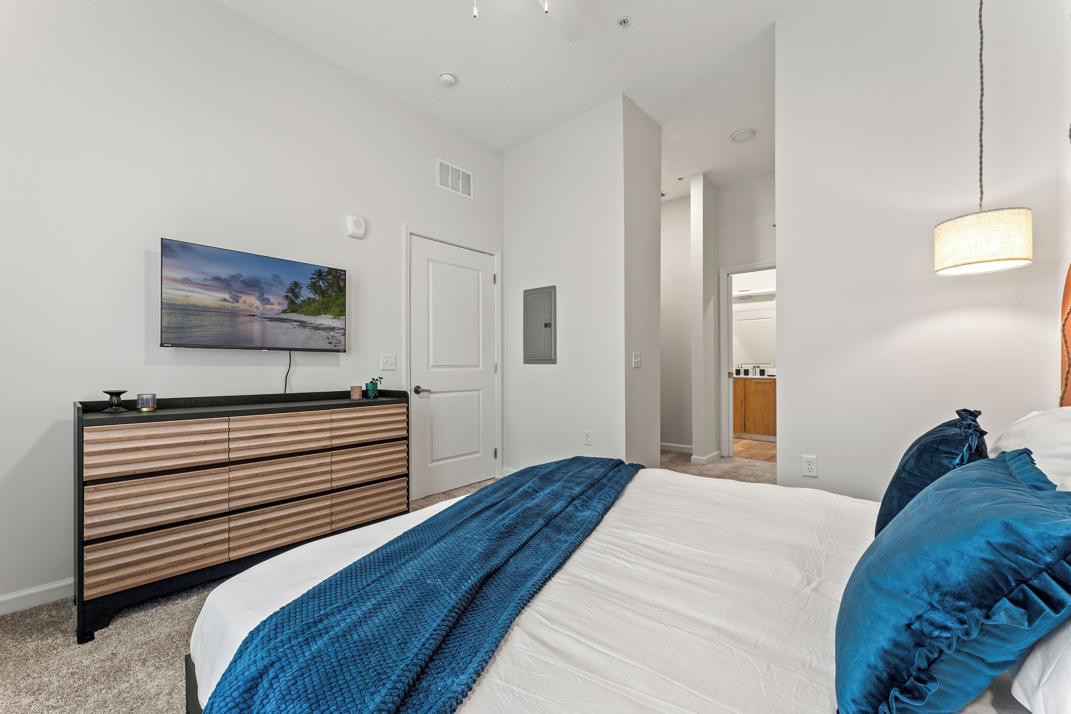 A bedroom with a bed, a dresser, a painting, and a hallway leading to a bathroom.