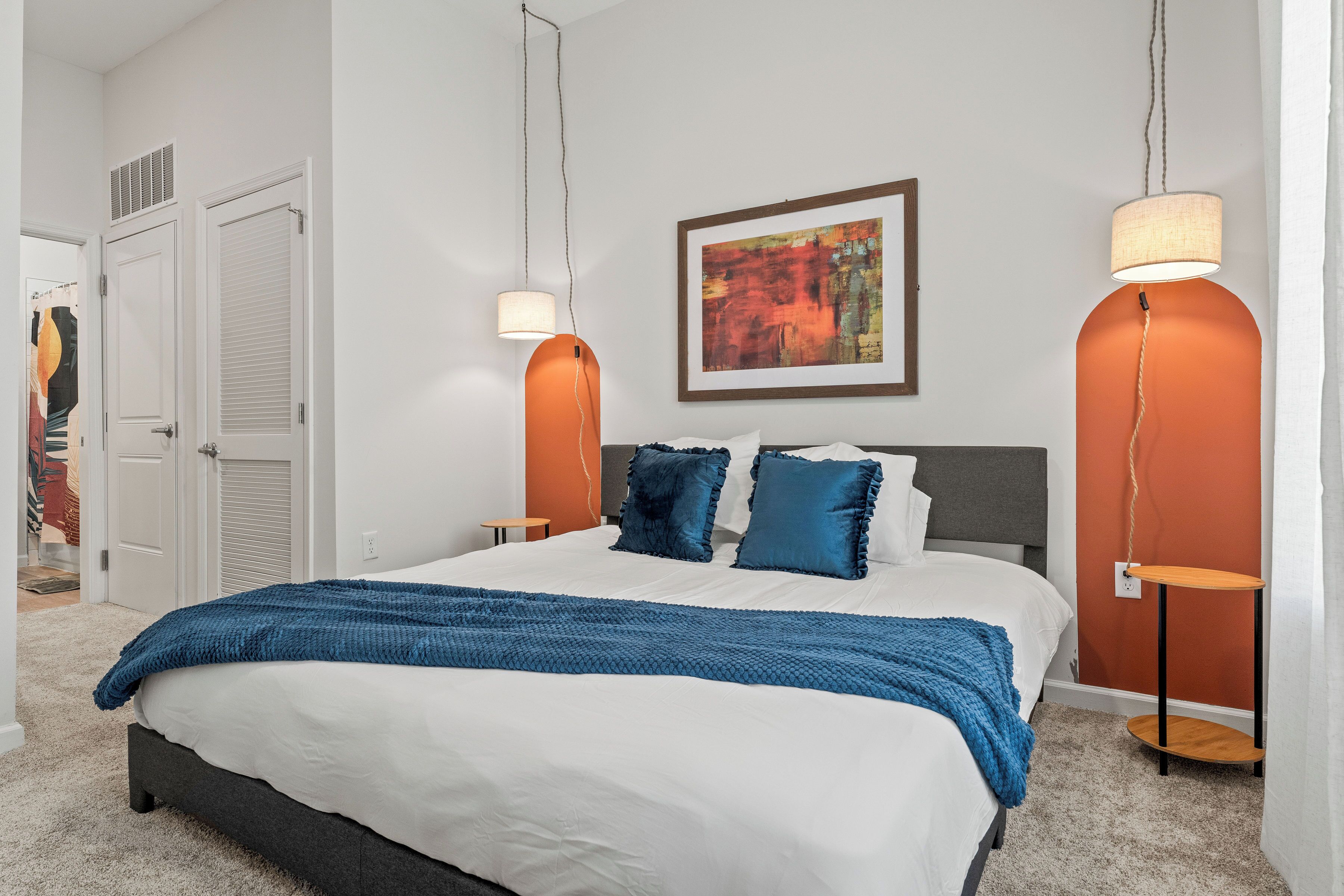 A bedroom with a large bed, two bedside tables, a framed artwork on the wall, and a door to the left.