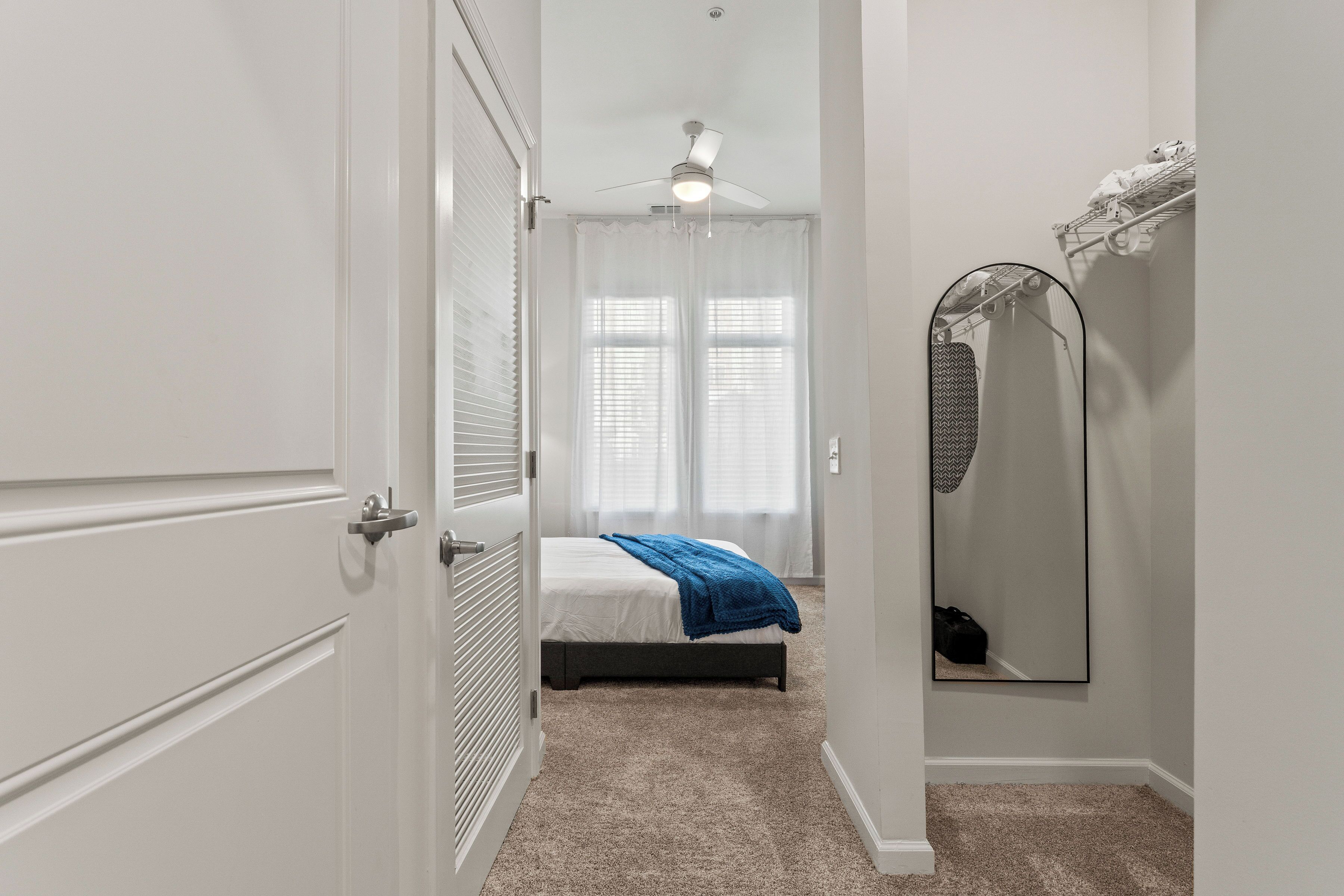 A bedroom with a bed, a ceiling fan, a window with blinds, a mirror, and a door.
