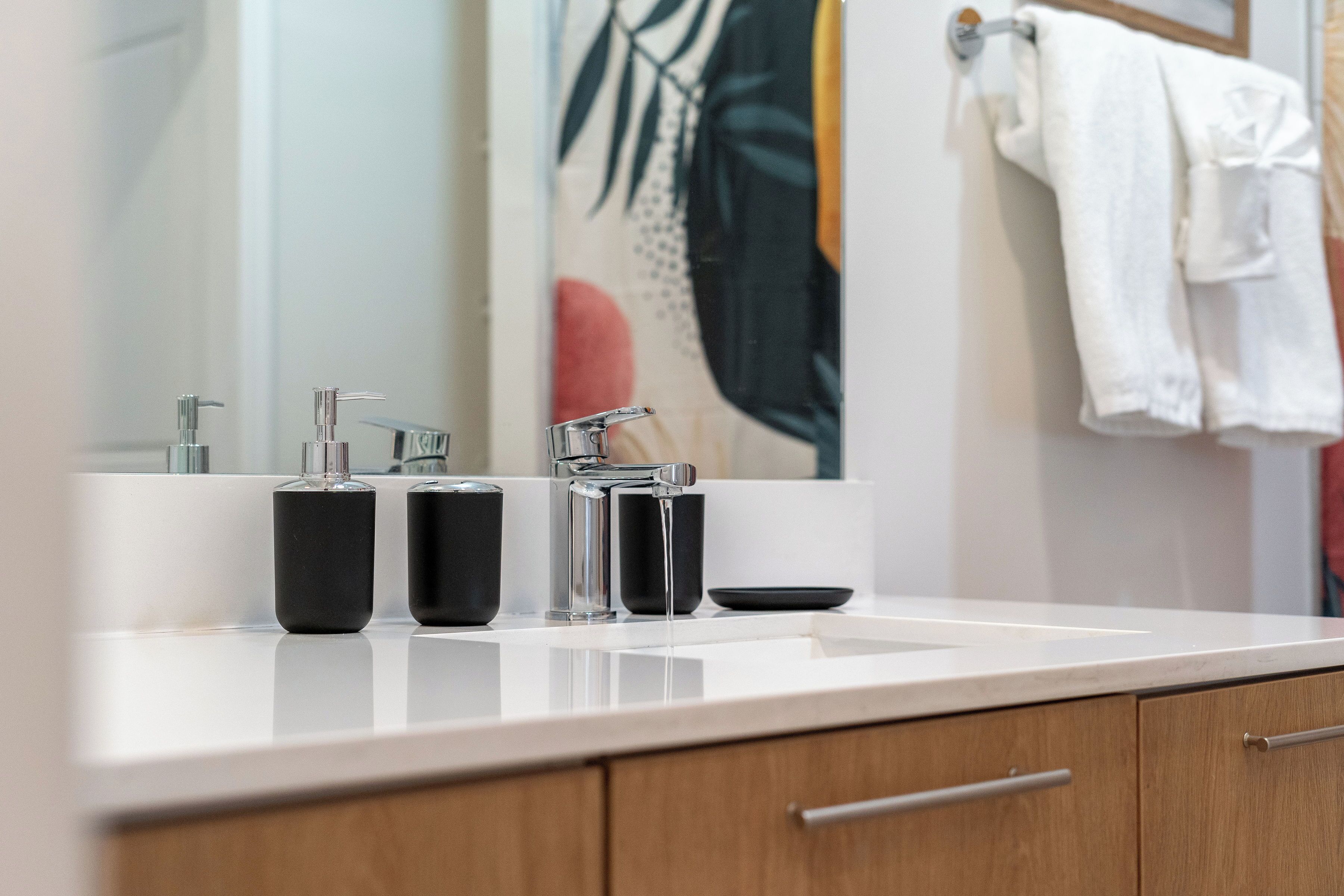 A bathroom with a white countertop, two black soap dispensers, a chrome faucet, and a wooden cabinet with a drawer.