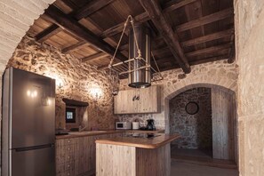 A rustic kitchen with a stone wall, wooden ceiling beams, a stainless steel refrigerator, wooden cabinets, and a hanging light fixture. - Vertigo Stone Villa - Roumeli Private Pool Escape (Mylopotamos)
