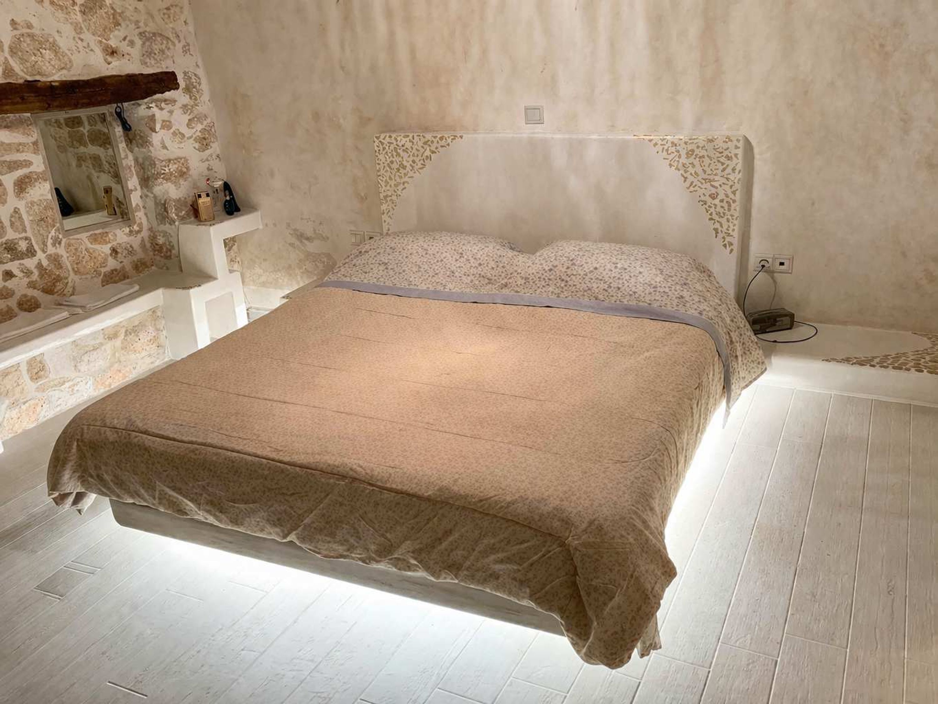 Een slaapkamer met een stenen muur, een houten balk, een wit bed met een gestreept hoofdeinde en een klein plankje met spullen.