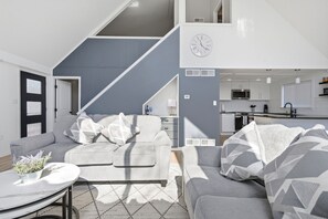 Un salon moderne avec un escalier, des canapés gris, un coin repas et une cuisine.
