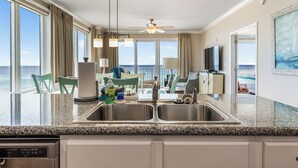 A kitchen with a double sink, a dishwasher, and a dining area with chairs and a table. - Gulf-Front Corner Unit with Stunning Panoramic Views (Panama City Beach)