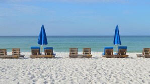 A beach with wooden lounge chairs and blue umbrellas. - Gulf-Front Corner Unit with Stunning Panoramic Views (Panama City Beach)