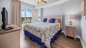A bedroom with a bed, a desk with a TV, a ceiling fan, and a nightstand with a lamp. - Gulf-Front Corner Unit with Stunning Panoramic Views (Panama City Beach)