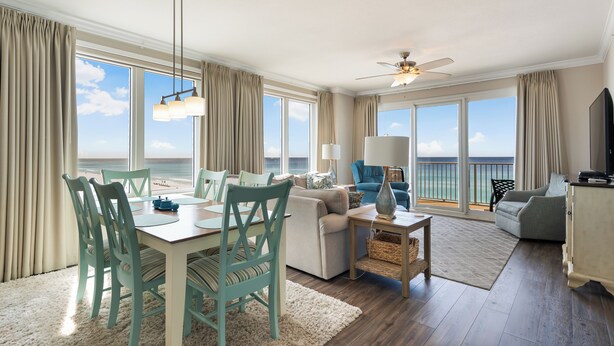 A spacious living area with a dining table, chairs, sofas, and a TV. Large windows offer a view of the ocean. - Gulf-Front Corner Unit with Stunning Panoramic Views (Panama City Beach)