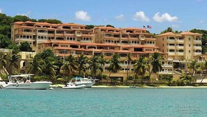 A beachfront hotel with multiple balconies, a flag, and several boats docked in the water. - Stunning Penthouse at Grande Bay - Breathtaking Views and Luxury Living! (St John)