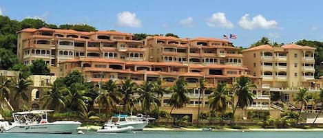 Un hotel frente a la playa con varios balcones, una bandera y varios barcos amarrados en el agua.